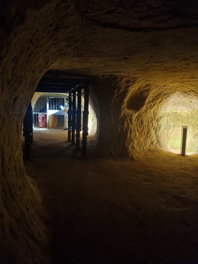 The image depicts a subterranean tunnel with rough, carved walls, likely composed of sandstone or a similar material. The tunnel walls are illuminated by an unknown light source above and at the end of the tunnel. A dark metal grate, supported by horizontal beams, spans the tunnel at a lower level. Behind the gate is a lighted area.  Red boxes or containers are visible. The tunnel appears to be a part of the Homburg Castle Caves, a historical sandstone mine located in Homburg, Germany.