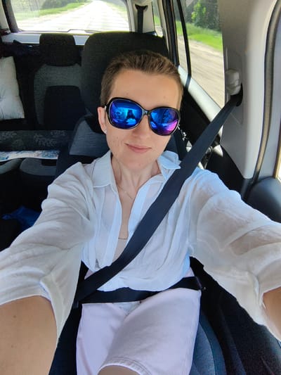 A person is taking a selfie in the front passenger seat of a car. They are wearing large, dark sunglasses with blue and pink reflections, a loose white button-down shirt, and light pink pants. The person has short, light brown hair and is wearing a delicate necklace and an earring. The car's interior is visible, including a black seatbelt across their chest and part of the back seat. The background outside the car window shows a road or path lined with green trees and vegetation, indicating an outdoor, potentially rural setting. The image appears to be taken during the daytime, with bright sunlight. There is no specific indication of the location being within Ilyanskiy selskiy Sovet, Belarus.