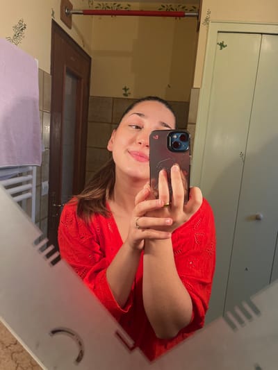 A female individual is depicted in a mirror selfie, centered from the upper chest upwards, within an indoor setting in Ancenis, France. She possesses dark hair pulled back and a smiling facial expression. She is wearing a red, long-sleeved top featuring eyelet embroidery. A gold-colored ring is visible on the ring finger of her left hand. She holds a dark-colored smartphone, equipped with a triple-lens camera array, in both hands, directing it towards the mirror for the photograph.
The reflected background reveals elements consistent with a bathroom or dressing area. To the left, a white heated towel rail is partially visible, draped with a light purple towel. Behind the subject, a dark brown wooden door with a translucent upper panel is present. Above the door, a red-accented horizontal bar is mounted on a light-colored wall. The walls are a light beige or cream shade, adorned with repetitive green stenciled botanical patterns. To the right, a light green cabinet or closet with two circular light-colored knobs is visible, also featuring similar green stenciled motifs. The mirror itself exhibits a frosted or etched geometric decorative pattern on its lower edges. The visible floor in the bottom left reflects as light-colored tiling.