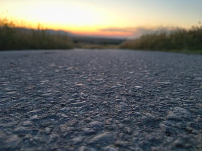 The image shows a low-angle, close-up view of an asphalt road in Razdelna, Bulgaria, at sunset. The foreground is dominated by the textured surface of the road, with visible stones and imperfections. On either side of the road, blurred vegetation extends into the distance. The horizon features a gradient of sunset colors, transitioning from bright yellow at the top to orange and then to a muted blue at the bottom. In the far distance, mountains are faintly visible against the colorful sky. The scene is devoid of any people or discernible actions.