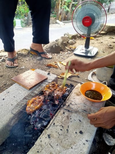 This image depicts an outdoor grilling scene in Celep, Indonesia. A makeshift charcoal grill, constructed from concrete blocks, occupies the center foreground. A metal grate holds two large, marinated pieces of meat, exhibiting charring and a glossy coating, cooking over glowing charcoal embers.
A person's right arm and hand are visible on the immediate right. This individual is actively basting the meat with a light green brush. An orange bowl containing a dark liquid sauce is positioned on the concrete surface beside the grill, from which the basting liquid is drawn. Metal tongs are held by this person's other hand, resting on the concrete. To the left of the grill, a rectangular wooden board is placed on the concrete surface.
In the middle ground, behind the grill, a red and black "MASPION" brand floor fan stands on a silver base, aimed towards the cooking area. To the left, the lower half of a second individual is visible, wearing black pants and black and grey flip-flops, standing on the dirt and gravel ground, which is scattered with dry leaves.
The background features a paved road. Beyond the road, green foliage, including banana trees, and a white structure are visible. A white chicken can be faintly discerned on the road in the far background.