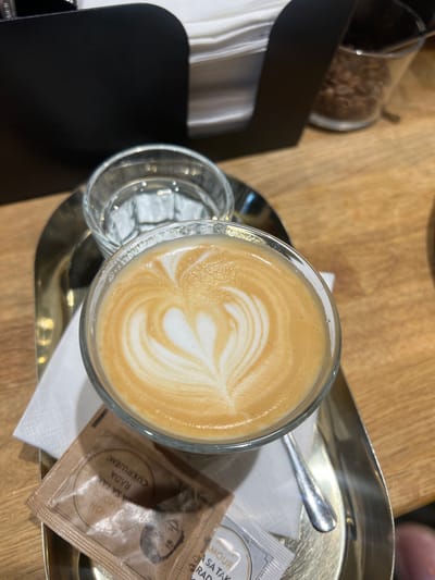 A close-up, eye-level shot shows a latte in a clear glass mug on a metal tray, positioned on a wooden table. The latte has a heart design in the foam. To the left of the latte is a glass of water. In front of the latte is a white napkin and a brown sugar packet with text in an unknown language. A silver spoon rests on the tray to the right of the latte. In the background, a black napkin dispenser holds white napkins, and a glass container filled with coffee beans is visible. The overall setting appears to be a cafe in Vysoké Tatry, Slovakia.