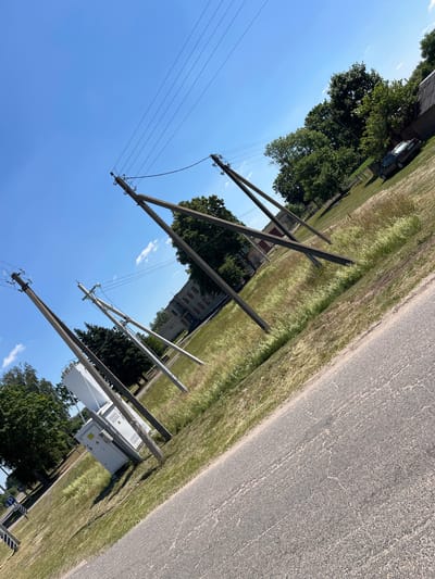 The image shows a rural road in the village of Astrumitski Sel'ski Savet, Belarus, on a clear, sunny day. Several power poles, some appearing old and leaning, are visible along the side of the road, supporting electrical wires. One power pole is accompanied by a large, white electrical transformer box. In the background, there are trees, a building, and a dark-colored car parked on grassy terrain. The road surface is asphalt, showing some wear and tear. The overall scene depicts a functional, yet slightly weathered, rural infrastructure.