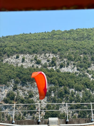 The image depicts an orange paraglider with a person attached, soaring in the sky. The paraglider is positioned in the foreground, partially obscuring a rugged, green-covered cliff face in the background. The cliff is densely populated with trees and shows significant rock formations. To the right of the cliff, a metallic structure, possibly a safety net or railing system, is visible, attached to a concrete surface. The sky is a clear, bright blue, suggesting good weather conditions, likely during the daytime given the lighting. A faint reddish-brown border is present on the left edge of the image, suggesting it was taken from within a structure or through a frame. There is some visible text on the paraglider wing that appears to read "FUSE". The overall scene suggests an outdoor recreational activity, likely paragliding, taking place in a mountainous or cliffside environment. The setting implies a location in Nances, France, known for such natural landscapes.