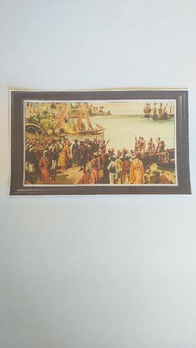 The image displays a photograph of a framed historical painting or print, presented against a plain white background. The painting depicts a busy port scene in the city of Limpok, Indonesia, characterized by a large gathering of people along the waterfront and numerous ships in the harbor.

The environment shows a body of water in the middle ground, bordered by land on both the left and right sides. On the left, lush green vegetation, including palm trees, and light-colored architectural structures are visible behind a crowded shoreline. On the right, a wooden pier or dock extends into the water. The sky above is a clear, light blue.

Numerous individuals are present, engaging in various activities. On the left side of the scene, a large crowd congregates by the water's edge. This group consists of diverse individuals dressed in a mix of traditional attire, including robes and turbans, and more formal European-style clothing. A central interaction is occurring where a figure in a prominent yellow/gold robe appears to be engaging with another group of people. Some figures carry what appear to be spears or muskets. Behind this group, a structure resembling a canopy or palanquin is discernible. On the right side of the image, a smaller group of people is active on the pier, some appearing to be partially clothed, possibly laborers.

In the water, various vessels are present. Smaller sailing boats with lateen sails are closer to the left shore, while larger, multi-masted sailing ships, possibly caravels or galleons with square sails, are positioned further out in the harbor on the right. The overall scene conveys a bustling maritime hub with significant human interaction and trade or diplomatic exchange.