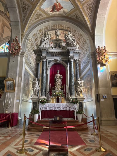 The image shows the interior of a religious building, likely a church or chapel, in Grad Rovinj, Croatia. The main subject is a highly ornate altar featuring marble columns, sculptures of religious figures, and decorative floral arrangements. A red cloth drapes behind the central statue. The ceiling is adorned with frescoes and decorative patterns. Below the altar, a red carpet is visible, bordered by stanchions with red ropes. A portion of a chandelier is seen hanging from the ceiling. The floor has a patterned tile design. Paintings are hung on the wall to the side of the altar.