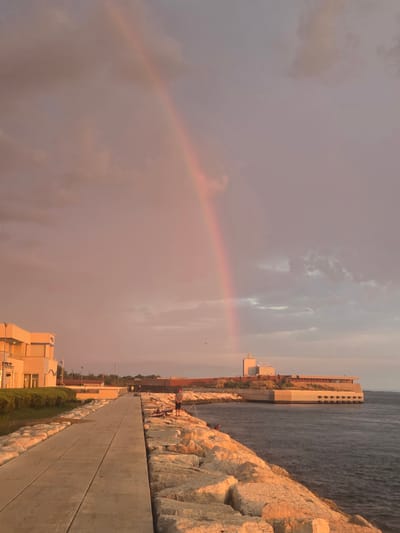 The image depicts a coastal scene in Chioggia, Italy, with a prominent rainbow arching across a muted sky. The foreground features a paved walkway bordering a rocky breakwater. Several individuals are present, some appearing to be fishing from the rocks. On the left side of the walkway, a modern building with cream-colored facades and blue-tinted windows is visible, bordered by manicured greenery. Further in the distance, along the coastline, another long, low-lying building with a contemporary design, featuring a brown roof and large windows, is situated. The sea, a deep blue, gently laps against the rocky shore. The overall lighting suggests either dawn or dusk, casting a warm, reddish-orange hue over the scene, particularly on the rocks and the sky.