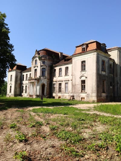 The image depicts a large, abandoned manor house in a rural setting, likely in the Жалудоцкі сельскі Савет, Belarus. The building is constructed of light-colored stone and has a multi-pitched roof with red tiles. Many of the windows are dark, suggesting the interior is unlit or empty. The facade exhibits signs of decay and neglect, with crumbling plaster and weathered stonework.
A grand, columned entrance with a damaged balcony is a central feature of the building's front. The surrounding grounds are overgrown with grass and weeds, and the foreground shows a dirt path with tire tracks, indicating recent activity or former access. A large tree with green foliage stands to the left of the manor, and a clear blue sky dominates the upper portion of the image. The overall atmosphere is one of desolation and historical abandonment.