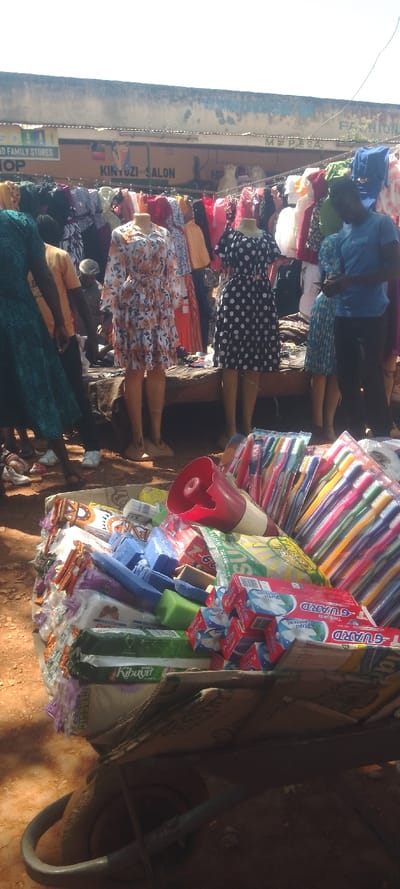 The image depicts an active outdoor market scene in Shirungu-Mugai ward, Kenya, under bright daylight. In the immediate foreground, a wheelbarrow is partially visible, functioning as a base for a retail display of various packaged goods. This display includes multiple bundles of colorful plastic toothbrushes, stacked boxes of "G-GUARD Pearls Whitening" toothpaste, individual blue and green soap bars, and plastic-wrapped items such as "Kibuyu" brand products and other toiletries. A red plastic megaphone with a white handle is positioned among these items.
The midground features a clothing stall with numerous dresses displayed on racks and three prominent headless female mannequins. The leftmost mannequin wears a white dress with an orange floral pattern, long sleeves, and a tiered skirt. The central mannequin displays a short-sleeved black dress with white polka dots. The rightmost mannequin wears a light blue or teal textured dress. Several individuals are present: a person in a dark green patterned dress is partially visible on the far left, an individual in an orange t-shirt is partially obscured behind them, and a male wearing a blue t-shirt and dark pants stands on the right, looking down at a mobile device. The ground is unpaved, reddish-brown soil.
The background shows a single-story commercial building with a faded blue-grey upper wall and a yellow-orange lower section, topped by a corrugated metal roof. Visible signs on the building include "AND FAMILY STORES," "KINTUZI SALON," "M-PESA," and a partial "FASHION" sign. A thin cable or power line runs across the upper portion of the background against a bright, clear sky.