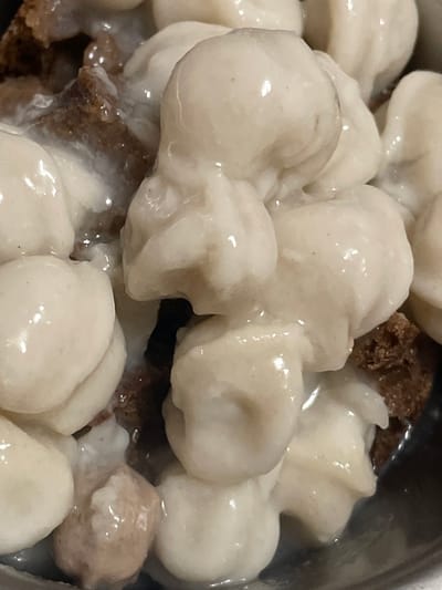 The image shows a close-up view of a bowl of food, likely a dessert or a dish with dumplings. The food consists of numerous small, rounded, white, and slightly translucent dumplings submerged in a creamy white sauce. Interspersed among the dumplings are pieces of dark brown, textured food, which appear to be bread or cake. The lighting is direct, highlighting the glossy surface of the dumplings and the sauce, with some reflections visible. The composition is tight, focusing entirely on the contents of the bowl, and no background or environment is visible. There is no indication of any specific location in Mirnyy, Russia, or any people, actions, or interactions.