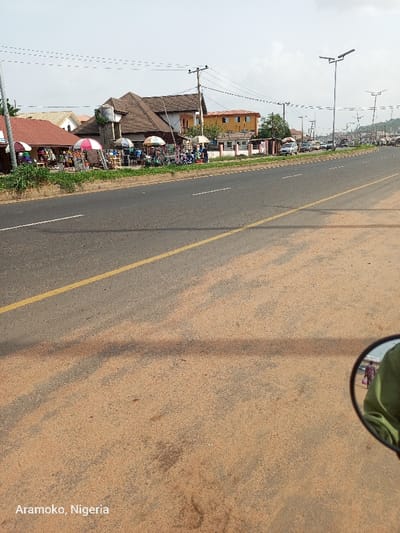 The image shows a view of a paved road in Ado Ekiti, Nigeria. The road has a single yellow line in the center and is mostly empty, with a few vehicles parked on the side. The road surface is covered in a thin layer of sand. There are several buildings on the left side of the road, and the buildings are all multi-story and made of concrete. There are also a few trees and bushes along the side of the road. The image is taken from the perspective of someone sitting on a motorcycle looking straight ahead, with a small rearview mirror visible in the lower right corner of the image. The mirror shows two figures in the distance. The image was taken on a clear day with some clouds in the sky. The location is a busy part of the city. The coordinates are 7.651 latitude and 5.226 longitude.