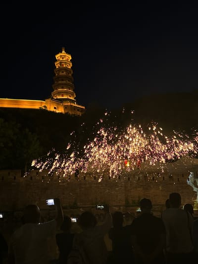 The image shows a night scene in Miyun District, China. A tall pagoda-like structure is illuminated on the left against a dark sky. To the right, a brick wall is lined with people taking photos, possibly of fireworks or a light display that appears as a cluster of bright lights. The scene suggests a public event or celebration is taking place.