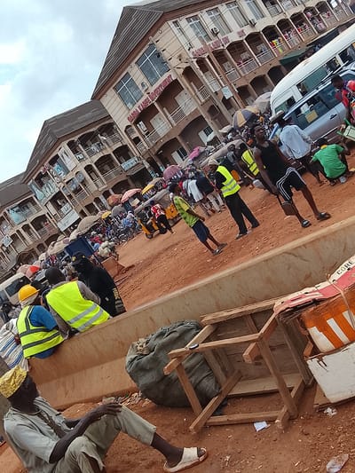 The image depicts a busy scene in what appears to be a commercial area in Katambe, Nigeria. In the foreground, an older man sits on the ground wearing a light-colored outfit and a yellow head covering. Beside him are a burlap sack, a simple wooden stool turned upside down, and cardboard boxes. Further back, construction workers wearing yellow vests and hard hats are sitting on a concrete barrier. The background is filled with a bustling crowd of people, many holding umbrellas. There are also parked vehicles including a van and some motorcycles. Buildings with multiple stories and storefronts are visible, including one with the sign "GARBA KADIBE INVESTMENT." The ground is a reddish-brown color.