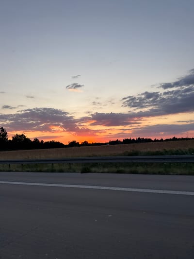 The image captures a sunset over a rural landscape near Malsch, Germany. The sky displays a gradient of soft blues and grays, transitioning to vibrant oranges and reds near the horizon, illuminated by the setting sun. Several clouds are scattered across the sky, some tinged with the warm hues of sunset. Below the sky, a line of trees forms a dark silhouette against the brighter sky, bordering a field of dry, golden grass or crops. In the foreground, a gray asphalt road or highway is visible, with a white line marking its edge. A metal guardrail separates the road from the grassy verge and the field beyond. The scene appears to be viewed from a moving vehicle, given the perspective of the road. No people or specific structures are clearly identifiable, creating a serene, naturalistic depiction of the countryside at dusk.