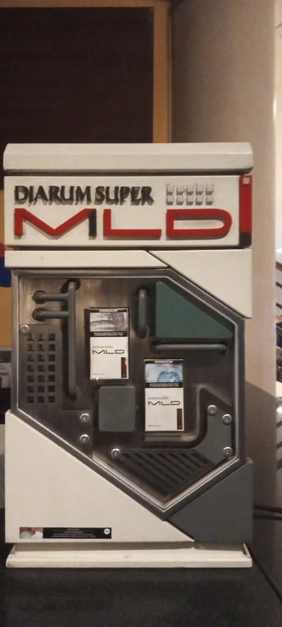 This image depicts a Djarum Super MLD cigarette display unit located indoors in the city of Aral, Kyrgyzstan. The unit, predominantly white with grey and red accents, features a prominent top panel with raised letters spelling "DIARUM SUPER MLD". Below this, a metallic-textured central panel displays two vertical packs of "DIARUM SUPER MLD" cigarettes. Each pack bears an Indonesian health warning ("PERINGATAN") and a graphic image. This central panel incorporates industrial design elements such as faux pipes, grilles, and visible rivets, alongside a dark green translucent trapezoidal insert and a small grey rectangular block. A small black label on the white base of the unit reiterates a "PERINGATAN" (warning) in Indonesian, includes an "18+" age restriction symbol, and displays a partial image of a person's face. The background consists of indistinct dark brown and light beige surfaces, consistent with an interior setting. No individuals are present in the scene.