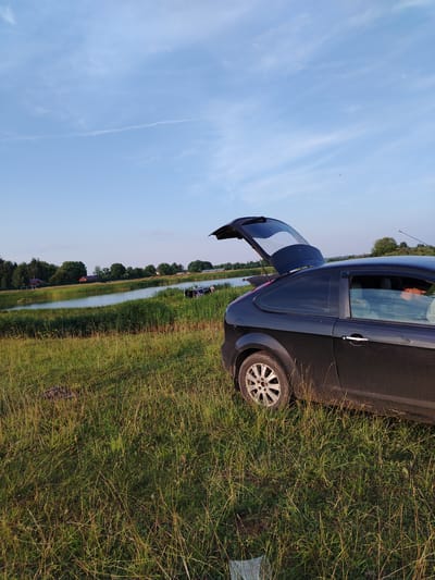 A black hatchback car with its rear hatch open is parked in a grassy field next to a body of water. The car is angled towards the right side of the frame, with its rear wheel clearly visible. A person is visible in the driver's seat. The water is surrounded by tall green reeds and further in the background, there are trees and houses. The sky is blue with some wispy clouds and a contrail from an aircraft. The scene appears to be in a rural area, possibly Kriukovskoe, Russia, based on the provided location context.