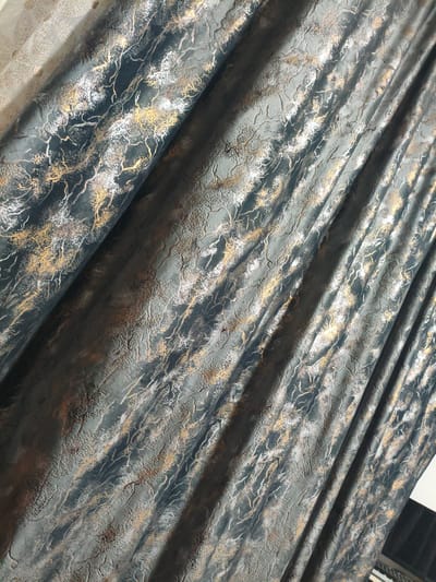 The image shows a close-up, angled view of dark gray curtains with a textured pattern that appears to resemble tree branches or veins. The pattern is highlighted with metallic gold and silver accents, creating a shimmering effect. The curtains are draped, forming vertical folds and shadows, which add depth and dimension to the fabric. The lighting suggests an indoor setting, possibly a room with natural light coming from the side. There are no visible people, animals, or distinct architectural elements that would indicate a specific location within the city of Luxembourg, Kyrgyzstan. The focus is entirely on the textile itself.
