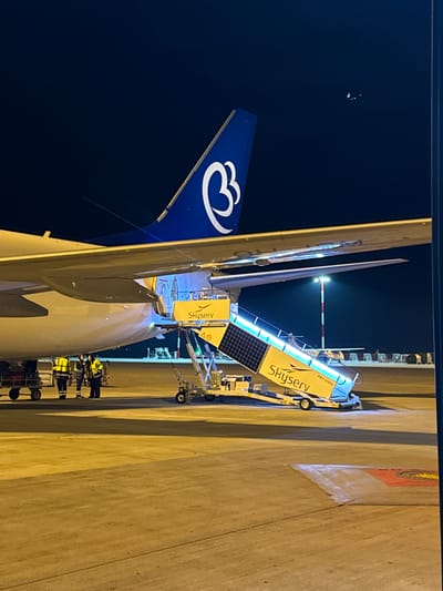 The image depicts an nighttime scene on the tarmac of Athens International Airport "Eleftherios Venizelos" in Paiania Municipal Unit, Greece. A large passenger aircraft, identifiable by its blue tail featuring a white stylized 'B' or wing-like logo (consistent with Aegean Airlines/Olympic Air), is parked on the illuminated concrete apron. A mobile passenger stair, branded "Skyserv" and labeled "PBT-31623," is positioned against an open aircraft door, illuminated by blue LED lights and equipped with visible solar panels. To the left of the stairs, approximately three ground crew personnel, wearing high-visibility yellow jackets and face masks, are gathered near the aircraft's fuselage, adjacent to two baggage carts or equipment trolleys. The background includes a tall, red and white striped airport light mast with an active light source, and distant airport infrastructure under a dark night sky.