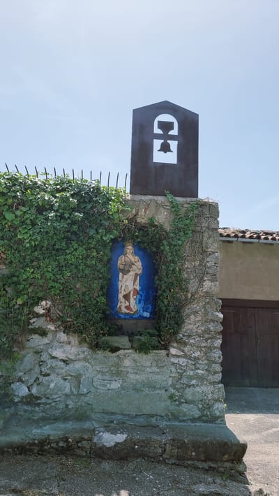 The image shows a rustic stone wall in the town of Susqueda, Spain, partially covered in dense green ivy. Embedded within an alcove in the wall is a vibrant blue tiled painting of a woman holding a baby, both wearing ornate crowns. The painting is framed by the rough texture of the stone and further softened by the creeping vines. Above the painting, a dark, angular silhouette of a bell tower is visible against a clear blue sky. The bell tower has a cutout in the shape of a bell. To the right of the stone wall, a building with a tiled roof and dark wooden doors is partially visible. The ground in front of the wall appears to be a mixture of gravel and dirt.