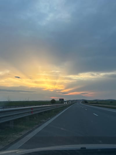 The image depicts a view from a vehicle moving on a highway near Plodovitovo, Bulgaria. The road is asphalt and features lane markings. There are guardrails along the side of the road with some red markings. Other vehicles are visible in the distance. The sky displays a dynamic mix of clouds, with the sun appearing to break through, creating a golden-orange glow.