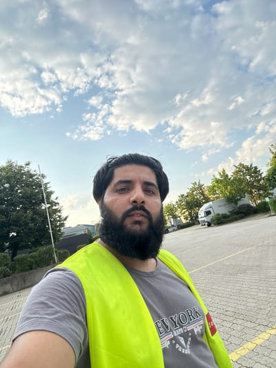 A selfie shows a man with dark hair and a full beard wearing a grey t-shirt and a bright yellow safety vest. The t-shirt has "NEW YORK" printed on it in red, white, and blue lettering. He is standing outdoors in what appears to be a parking lot, possibly at a truck stop or industrial area. In the background, there are trees, buildings, and several trucks, including a white Mercedes-Benz truck. The sky is partially cloudy. This image was captured in Schweitenkirchen, Germany.