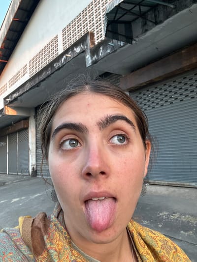 The image depicts a close-up, eye-level view of a single individual, a female with dark hair, green eyes, and fair skin, performing an action. She is looking upwards and outwards, with her mouth open and tongue extended fully. Her hair shows visible gray strands at the part on the top of her head, and a small red blemish is present on her forehead. She is wearing a patterned garment, predominantly yellow and brown with interwoven colors, and a silver-colored filigree earring is visible on her right earlobe.
The background places the scene in an urban environment consistent with Chiang Rai City Municipality, Thailand. Behind the individual, a low-rise building structure is visible under natural lighting. The building features light-colored, possibly concrete, exterior walls with horizontal sections of square ventilation blocks. A weathered, dark concrete ledge or overhang runs horizontally across the mid-section of the building. Below this ledge, multiple grey metal roller shutter doors, typical of commercial storefronts or storage units, are visible in a closed position. The architectural style, including the concrete construction, ventilation blocks, and roller shutters, is characteristic of urban areas in Thailand. Partial views of a sloped roof with corrugated material and some visible structural elements or piping are present above the building's facade. The ground appears to be a paved surface.