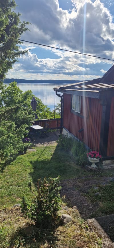 The image depicts a waterside view of a red wooden house in Sigtuna, Sweden. The house has a dark roof and a visible window. In front of the house, there is a green lawn with a small bush and some rocks. To the left of the house, a wooden deck with a table and chairs is visible, partially obscured by trees. A large body of water stretches across the horizon, with a forested area on the far bank. The sky is filled with dramatic, sunlit clouds. The overall scene suggests a serene, natural setting.