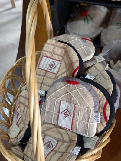 A woven basket overflows with several light brown, quilted bags, each featuring a red embroidered symbol on a white and red patterned patch. The bags are circular or dome-shaped and have stitched patterns resembling a grid. Some bags have contrasting colored edges in black and gray. Several small tags with the words "FELT WORLD" are visible on the bags. In the background, other similar items, possibly slippers, are stacked, one with a red flower embroidery. The setting appears to be a shop or market stall in Кызыл-Туу, Kyrgyzstan, with a wooden surface visible beneath the basket.