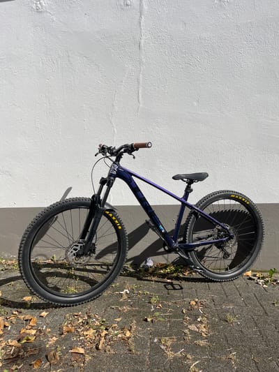 The media file shows a purple mountain bike leaning against a textured white wall. The bike is positioned vertically, with its wheels facing the right side of the frame, which is paved with dark grey asphalt interspersed with fallen leaves and sparse greenery. Sunlight casts a shadow of the bike onto the wall and the ground, indicating a sunny day.
The bike itself is a prominent object, with visible components like the saddle, handlebars, frame, gears, and tires. The tires are marked with "MAXXIS" in yellow. A small GoPro camera is attached to the frame.
The scene appears to be outdoors, likely on a street or pathway, given the pavement and fallen leaves. The lighting suggests it's daytime, possibly late morning or afternoon due to the angle of the shadows. No people are present in the image. The overall impression is one of a parked bicycle in an urban or suburban outdoor setting.
