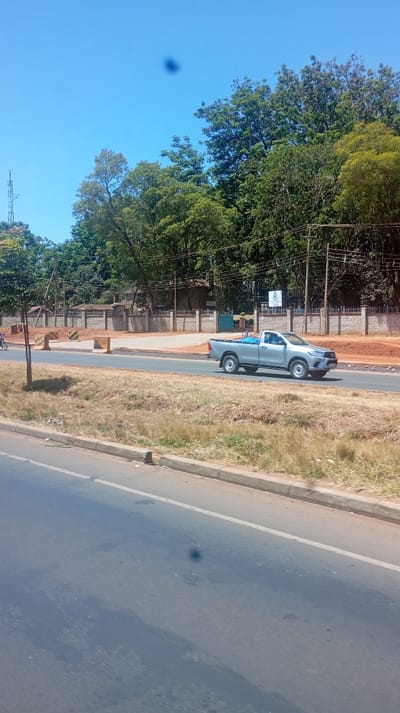 A silver pickup truck drives along a road on a clear day. A barrier or wall construction zone appears behind the truck. Trees are visible behind the wall, against a bright blue sky. The road curves to the left in the foreground. Other trees and an electrical tower are visible on the left side of the frame.
