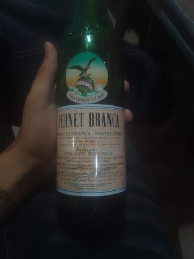 The image shows a green glass bottle of Fernet Branca. The label features an eagle perched atop a globe with "Branca" inscribed on it, and the words "Casa Fondata Nel 1845" beneath. "Fernet Branca" is prominently displayed, along with "Fratelli Branca - Distillerie." Additional text on the label mentions "Vicino alla Chiesa Dos" and "del famose Fernet Branca." The bottle is being held in someone's hand, with a dark background visible.