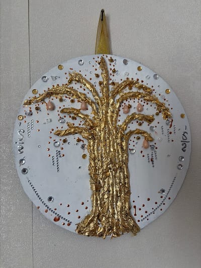 The image displays a circular wall art piece against a textured, light-colored wall. The artwork features a prominent golden tree in the center, with its trunk and branches textured and appearing to be made of crumpled gold leaf. Scattered around the tree are various embellishments, including small pink teardrop-shaped beads, clear and silver rhinestones, small gold and copper beads, and tiny gold sequins. The perimeter of the circular artwork is decorated with rows of small silver beads and copper dots, with some silver lettering visible on the left and right sides. A gold ribbon is attached to the top center of the artwork, suggesting it is meant to be hung. The overall impression is of a decorative, handmade piece of art. There is no indication of the city of Мяндиги, Russia within the image itself.