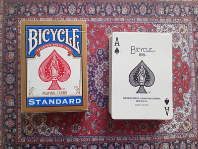 The image displays a deck of Bicycle brand playing cards, specifically a Standard deck box and a single card from a Bicycle 808 deck. The playing cards and their box are placed on a red oriental rug with intricate patterns in shades of blue, white, and dark red. The deck box is gold with blue and red lettering and features the Bicycle logo and the text "TRUSTED SINCE 1885" and "PLAYING CARDS STANDARD". The single card is an Ace of Spades from a Bicycle 808 deck, also showing the Bicycle logo and "808". Text below the spade reads "THE UNITED STATES PLAYING CARD COMPANY MADE IN U.S.A. 1025-F1612H". There are no people or any identifiable landmarks of the city of Bitburg, Germany, in the image. The scene is static and appears to be a still life arrangement.