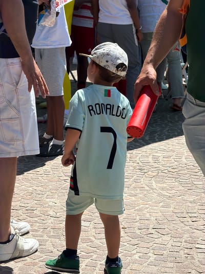 A child wearing a light blue soccer jersey with the name "RONALDO" and the number 7 on the back, along with matching shorts, is standing outdoors on a paved surface of beige and brown stones. He is also wearing a hat with a light-colored pattern. An adult's arm holds a red cylindrical object near the child. Other people are present, with parts of their clothing and bodies visible in the background. The environment appears to be a public space. There is no definitive information to identify the location as Fentange, Luxembourg, so this detail cannot be forensically confirmed from the visual evidence.
