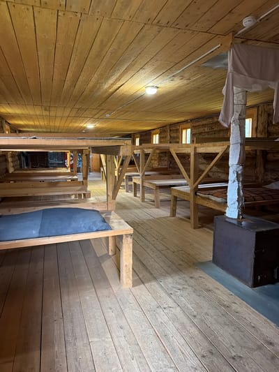 The image shows the interior of a rustic wooden dormitory located in Ust'-Pyzas, Russia. The room is furnished with multiple wooden bunk beds and single beds, some with mattresses and bedding. The walls and ceiling are constructed from natural wood planks. There are small windows letting in natural light. In the foreground, a dark blue mattress lies on a lower bunk. To the right, a dark, rectangular object, possibly a storage chest or heater, stands near a pillar covered with fabric. The flooring is made of weathered wooden planks. The overall impression is of a simple, communal living space with a strong emphasis on natural materials.