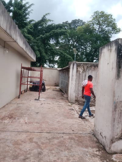 This media file captures an outdoor scene in Onibudo, Nigeria, featuring a man walking away from the camera. He is wearing a bright orange t-shirt, blue jeans, and brown sandals, with a light-colored cloth draped over his left shoulder. The man is walking on a worn, concrete ground with visible cracks and stains, suggesting a somewhat aged or weathered environment.
To the left of the frame, there's a red metal scaffolding structure. Behind it, large black plastic bags are piled up, hinting at construction, renovation, or perhaps waste management. The background is dominated by lush green trees with dense foliage, reaching towards a cloudy sky. A contemporary street lamp is visible amongst the trees, suggesting this might be an urban or semi-urban area.
The walls surrounding the area are made of concrete, showing signs of age with discoloration and staining, particularly on the right-hand wall which has dark patches. The overall atmosphere appears to be daytime, with diffused light due to the overcast sky. The man's posture and stride do not convey any specific emotion, presenting a neutral moment. There is no visible text in the image.