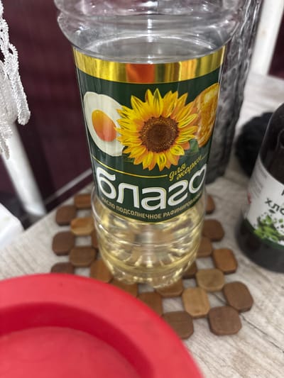 This media file displays a close-up, top-down view of a bottle of sunflower oil, with its label prominently featured. The bottle is made of clear plastic and contains light yellow oil. The label has a dark green background with a golden border. It features a large, detailed illustration of a sunflower with a fried egg next to it. Large white Cyrillic lettering on the label reads "СОЛНЦЕ" (Solntse), meaning "Sun" in Russian. Below this, smaller Cyrillic text reads "МАСЛО ПОДСОЛНЕЧНОЕ РАФИНИРОВАННОЕ" (Maslo podsolnechnoye rafinirannoye), which translates to "Sunflower oil, refined."
The bottle is lying on a surface that appears to be a wooden trivet composed of small, rectangular wooden pieces. To the right of the trivet, a bright red object, possibly a bowl or lid, is partially visible. In the background, another dark bottle, possibly containing a dark liquid, is out of focus. A white, lace-like fabric is visible in the lower portion of the image.
The scene appears to be indoors, on a table. There are no people or animals visible, and no clear indication of the time of day or weather conditions. The overall impression is a still life arrangement focusing on the product. The location context is Княгинино, Russia.