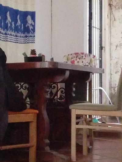 The image shows a close-up view of a table and chairs in what appears to be an indoor setting. A wooden table with intricate metalwork on its legs is visible, topped with a decorative cloth with a floral pattern. On the table, there are a few small objects, including what looks like a pine cone in a dark container. To the left of the table, a dark fabric object, possibly clothing, is draped over a wooden stool. To the right, a light-colored wooden chair with a green upholstered seat is partially visible. In the background, a window with vertical bars is present, and part of a white wall with textured or peeling paint is visible. The overall scene suggests a rustic or vintage aesthetic. The location is specified as Atamo Sur, Venezuela.