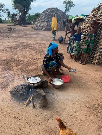An outdoor scene in Nandu, Nigeria, depicts a village setting. In the foreground, a woman wearing a blue headscarf and a patterned dress crouches next to a cooking fire. A metal pot sits on rocks over the fire, with smoke rising. She is holding a small metal bowl and a larger metal bowl containing a white liquid, with a red bowl nearby. Several children are visible in the midground, some standing near a thatched structure and others in the background. One child wears a yellow garment. The ground is sandy and dry, with several thatched huts and trees scattered throughout the landscape. A chicken is present in the lower right corner of the image.