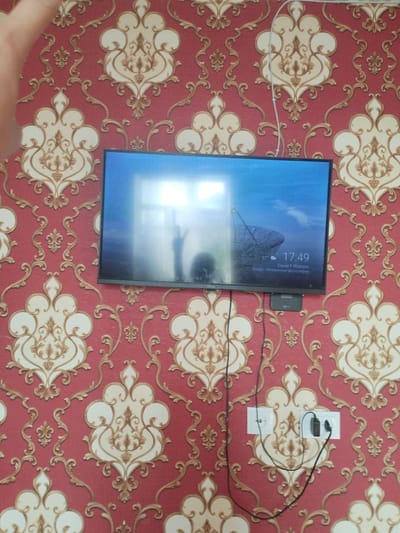 A television is mounted on a red and gold damask wallpapered wall in what appears to be a residential setting. The TV screen displays a satellite dish against a blue sky, with the time "17:49" and the name "David F Watson" superimposed. A small black device is attached to the bottom center of the TV, and wires run down the wall to two electrical outlets. One outlet has a black plug inserted, and the other is empty. The overall impression is that of a living room or bedroom. There is no visible indication of the location being in Mady, Kyrgyzstan, as this information is not present in the visual data.