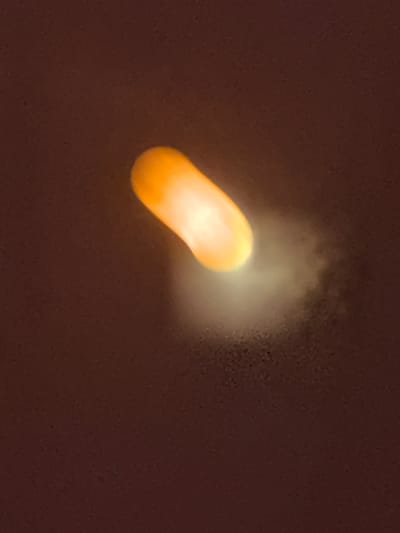 An oval, glowing orange object is suspended in the center of the frame against a dark, textured background. The object emits a soft, diffused light, creating a halo-like effect that extends outwards. The background appears to be a solid, uniform surface with subtle speckling. The overall impression is one of isolation and illumination. No specific location in Baryshevo, Russia, can be determined from the visual information provided.