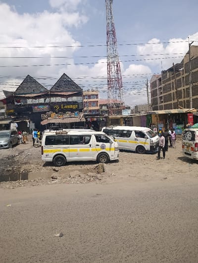 The image displays an outdoor urban scene in Matungulu west ward, Kenya, under a partly cloudy sky with scattered white cumulus clouds. The foreground consists of a paved asphalt road exhibiting minor debris. The midground features an unpaved, uneven dirt and gravel area that appears to function as a parking or waiting zone. Three white public transport minivans with yellow stripes are visible; two are prominently parked on the unpaved surface, and a third is partially visible on the far right. A silver sedan is partially visible on the far left. Approximately 10-15 individuals, predominantly adult males in casual attire, are present, standing or walking near the vehicles and buildings. In the left-to-center background, a multi-story commercial building with two dark, pyramidal roofs and a prominent "KS Lounge" sign on its facade is visible. To the right of this building, a tall, red and white lattice communications tower dominates the skyline. Other multi-story commercial and residential structures, some appearing incomplete with exposed construction elements, are visible behind and to the right of the primary commercial building. Overhead utility lines traverse the upper portion of the frame.