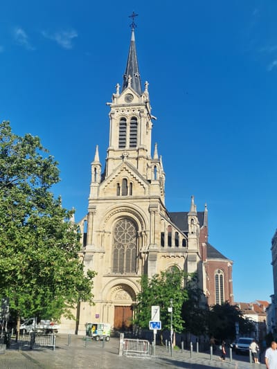 The image shows the Église Saint-Gilles in Saint-Gilles, Brussels, Belgium, under a clear blue sky. The church, built of light-colored stone, features a tall, pointed steeple with a clock face near its top. The main facade is dominated by a large rose window above an arched entrance. To the left, a large, leafy green tree partially obscures the church. In the foreground, a paved street area is visible, with several people walking and standing. A white truck and a small green utility vehicle are parked on the street. Various street signs and bollards are also present. The overall scene captures a sunny day in the urban environment of Saint-Gilles.