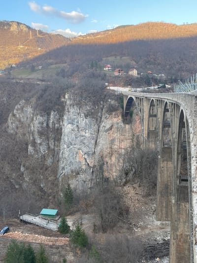 The image displays the Đurđevića Tara Bridge, a multi-arch concrete structure, spanning a deep river gorge in Đurđevića Tara, Montenegro. The bridge extends from the right foreground into the upper center, showcasing multiple supporting arches. The gorge walls consist of steep, rocky cliffs with varied grey and reddish-brown hues, sparsely covered by leafless deciduous trees and scattered evergreen conifers. At the base of the gorge on the left, a wooden platform supports a small building with a green roof, beside which a dark-colored sedan is parked on a dirt patch. Beyond the gorge, forested mountains rise, with the upper slopes of the mountain on the left receiving direct sunlight, casting an orange hue, while other slopes remain in shadow. Several small residential buildings with red roofs are visible on the distant hillsides. The sky above is clear blue with a few scattered white clouds. No individuals or specific actions are discernible within the frame.