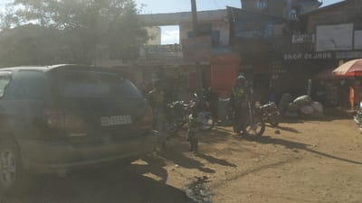 The scene is in Kayna, Democratic Republic of the Congo, at latitude -0.561, longitude 29.181. It appears to be a street in front of shops. A dark gray SUV is parked in the street with the back end facing the viewer. The vehicle has a license plate that reads "G7 864A 39." There are two motorcycles parked in front of the vehicle, and another motorcycle is parked in the distance. There are multiple people in the scene, though only one can be clearly seen - a man wearing a white helmet and a blue shirt is standing near the motorcycles. Several large, dark sacks are stacked on the ground next to the motorcycle in the distance. There is a shop sign with the words "Shop of Orange" on the facade of a building. An orange and white awning partially covers the sidewalk in front of the shop. The ground is a mixture of dirt and gravel. The sky is clear and blue. The scene is bright and sunny.