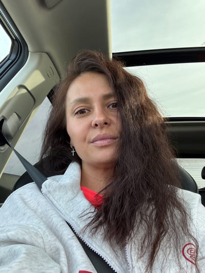 A woman with long, dark brown wavy hair and light brown eyes is seated in the driver's or passenger's seat of a vehicle, taking a selfie. She has full lips adorned with light pink lipstick and maintains a neutral, calm expression while looking directly at the camera. She is wearing a light grey zippered hoodie over a red top. A small silver cross earring is visible on her left ear. The grey hoodie features a stylized red logo on the right side of the chest. A black seatbelt is fastened across her.

The interior of the vehicle is visible, including a black car seat, a black seatbelt, and beige/grey trim on the door panel and roof lining. Bright, uniform light enters through the car window, indicating it is daytime, possibly an overcast day. The text "AIRBAG" is visible on the car's roof pillar. The scene takes place in Minderevo, Russia.