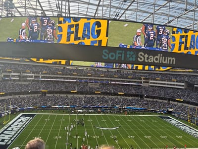 An image displays an American football game inside SoFi Stadium. The field is visible with teams positioned on the green turf. Above the field, a large screen shows a replay of the game. The screen displays the text "FLAG!" in a stylized font alongside images of players and referees. The stadium is filled with spectators. The "SoFi Stadium" logo is prominently displayed on a dark banner below the screen.