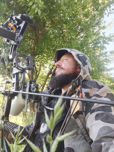 A man with a beard and mustache, wearing a camouflage hooded jacket and a black vest, is shown from the chest up, holding a compound bow. He is outdoors, with green foliage and trees in the background, suggesting a natural environment. The man is looking upwards and to the right, with a slight smile. The compound bow is partially visible in the foreground, with arrows attached. The lighting suggests it is daytime, possibly morning or late afternoon, given the angle of the sun. The location is Semypolky, Ukraine.