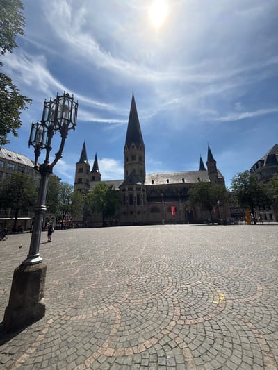 The image captures the Bonn Minster, a prominent landmark in Bonn, Germany, under a bright, sunny sky with scattered clouds. The cathedral, a Romanesque basilica, dominates the background with its imposing stone architecture and tall spires. In the foreground, a cobblestone plaza features a decorative pattern of circular stone arrangements. A vintage-style lamppost stands to the left, casting a shadow on the plaza. Several trees provide greenery on the sides of the plaza, and a few buildings line the distant street. The scene is bustling with a few people casually walking or standing in the plaza. A pigeon is visible on the cobblestones. The overall atmosphere is serene and picturesque, showcasing a sunny day in the historic city center.