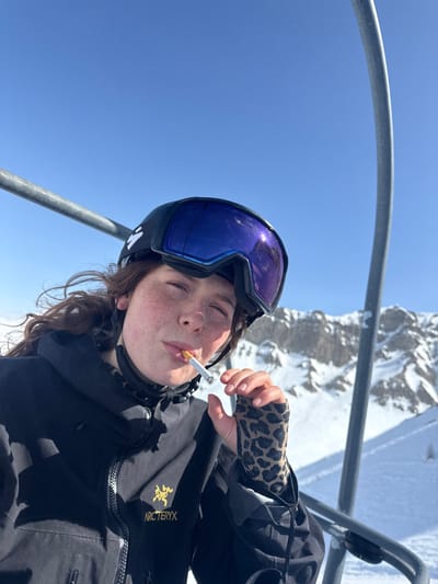 A female individual is photographed in a close-up, chest-up view, seated on a ski lift in Le Monêtier-les-Bains, France. She faces the camera with a slight upward gaze. Her head is covered by a black ski helmet with an unidentified white logo on the left side, and reflective blue-purple ski goggles are pushed up on the helmet. Dark, wavy hair is visible below the helmet. Her face shows freckles across the nose and cheeks. She wears a black ski jacket, identified by a yellow Arc'teryx logo on the left chest, and a leopard-print fingerless glove on her right hand. A lit cigarette is held between her pursed lips. The background consists of a clear blue sky and snow-covered mountain ranges under direct sunlight. Two gray metal ski lift poles are visible, framing the subject on the left and right sides of the image.